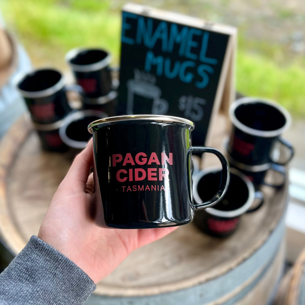 A hand holding a black enamel mug with 'PAGAN CIDER TASMANIA' text on it, displayed in front of a sign that reads 'ENAMEL MUGS'.