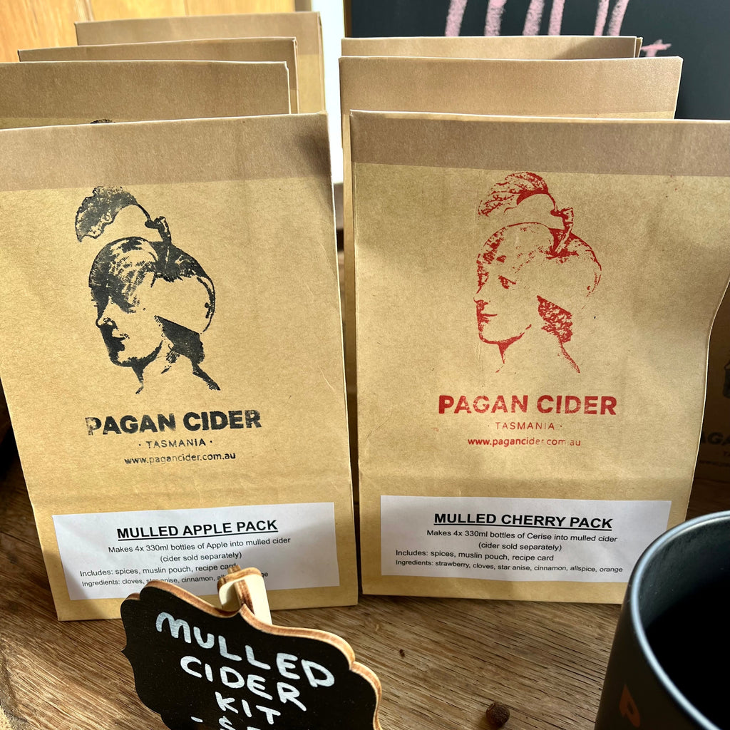 Several packets of Pagan Cider Mulled Apple and Mulled Cherry packs displayed on a wooden surface, with a small blackboard sign reading 'Mulled Cider Kit'.